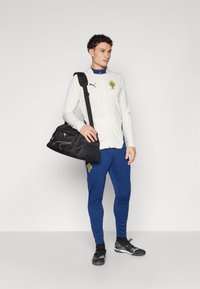 Giacca bianca con zip e dettagli blu, con logo. Pantaloni sportivi blu e borsa da palestra nera con tracolla. Scarpe da ginnastica nere con dettagli bianchi.