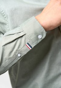 Camicia verde chiaro con bottoni e maniche lunghe. Presenta bottoni bianchi e un piccolo ricamo rosso, bianco e blu sul polsino.