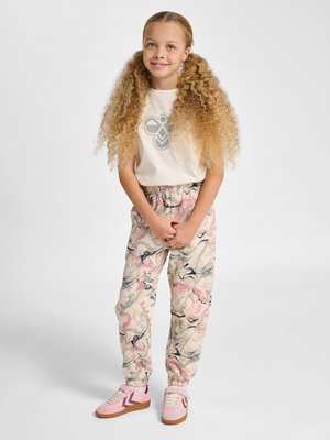 Young girl with long curly hair wearing white logo T-shirt, patterned jogger pants with pink and navy accents, and pink sneakers, standing and smiling.