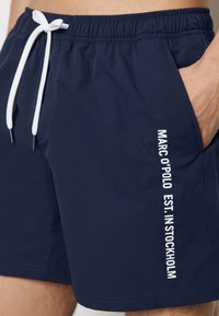 Costumi da bagno blu navy con coulisse bianca, vita elastica, tasche laterali e testo di branding verticale sulla gamba sinistra. Texture liscia.