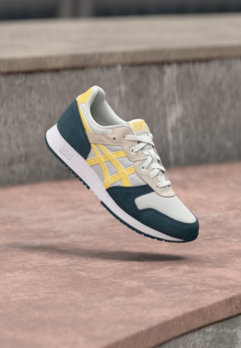 Beige og teal sneakers med gule accenter, mesh- og suedematerialer, klassisk snøring, teksturert såle, med ASICS-logo.