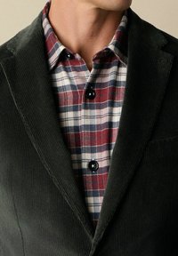 Ciemnozielona marynarka z korduroju nałożona na koszulę w kratę w czerwono-niebiesko-białe paski z ciemnymi guzikami. Posiada wyraźną fakturę i skośny kołnierz.