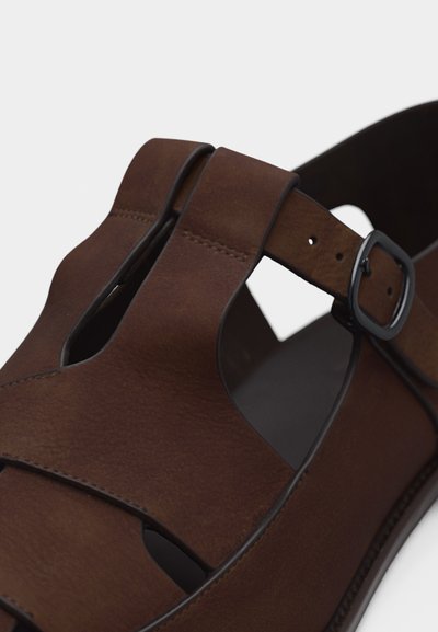 Καφέ δερμάτινο σανδάλι με ανοίγματα, μαύρο λουράκι με αγκράφα και εμφανείς ραφές σε λευκό φόντο.