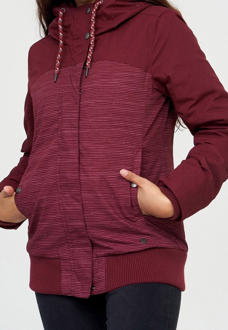 Femme portant une veste rayée bordeaux avec capuche et cordons, mains dans les poches, sur fond blanc uni.