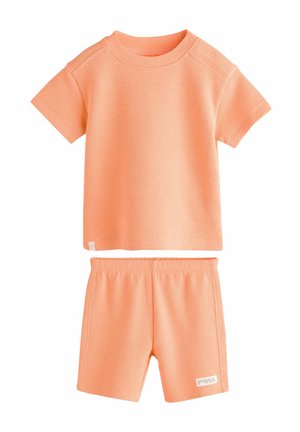 Maglietta a coste color pesco con maniche corte e bottoni a pressione sulla spalla, abbinata a pantaloni corti coordinati con elastico in vita.