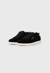 Baskets en suede noir au profil bas, avec des lacets noirs, une semelle en caoutchouc crème, et un logo brodé en blanc sur le côté.