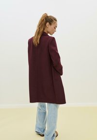Manteau en mélange de laine bordeaux au design ajusté, à manches longues et avec poches latérales, associé à un jean à jambes larges bleu clair.
