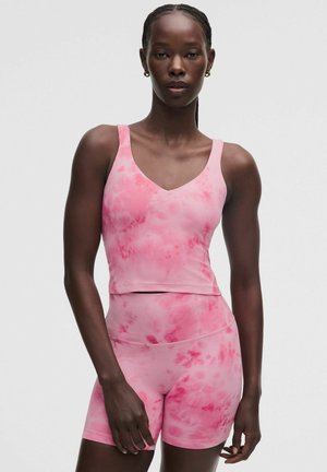 Szczupła osoba ubrana w różowy batikowany top bez rękawów i krótkie spodenki z wysokim stanem, stojąca na jednolitym jasnym tle.