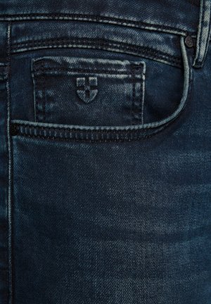Ciemnoniebieskie dżinsy z kieszenią na monety, w której wyhaftowany jest mały logo w kształcie tarczy.