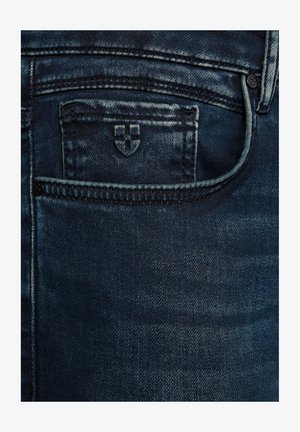 Ciemnoniebieskie dżinsy z kieszenią na monety, w której wyhaftowany jest mały logo w kształcie tarczy.