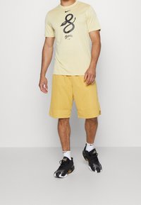 Camiseta Nike de color amarillo claro con un diseño gráfico negro, combinada con pantalones cortos amarillos hasta la rodilla. Usada con zapatillas negras y blancas.
