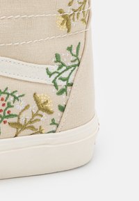 Sapatilha de tela bege com bordado floral em verde e amarelo. Possui sola de borracha branca e detalhes em tecido texturizado. Acentos de design simples.