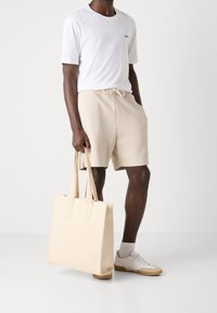 T-shirt blanche avec un petit logo, shorts beiges texturés et un sac tote léger beige. Le modèle porte des baskets blanches avec des accents beiges.