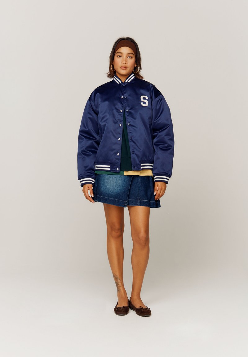 Veste varsity en satin bleu marine avec col et poignets rayés blancs, arborant une lettre "S" sur la poitrine, associée à une jupe en denim bleu.