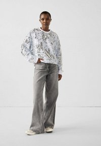 Persoon draagt een witte trui met pailletten, grijze jeans met brede pijpen en witte sneakers, staat met één hand in de zak tegen een effen achtergrond.