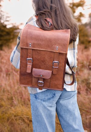 Brun læder rygsæk med klaplukning, metalhardware, to justerbare stropper og en frontlomme, sat mod en naturlig baggrund.