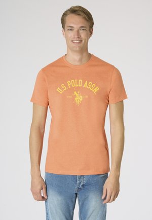 Giovane uomo sorridente, indossa una maglietta arancione U.S. Polo Assn. con logo giallo e jeans azzurri, in piedi su uno sfondo chiaro e uniforme.