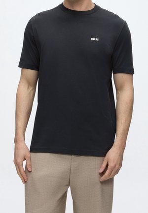 Mann trägt ein schwarzes T-Shirt mit kurzen Ärmeln und kleinem weißem "BOSS"-Logo auf der Brust sowie beige Hosen vor schlichtem Hintergrund.