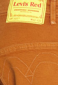 Braune Denim-Hosen mit einem gelben Etikett mit der Aufschrift "Levi's Red." Auffällige Nähte in einem geschwungenen Muster auf der Gesäßtasche. Größe W32 L32.
