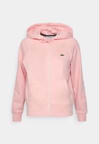 Světle růžová mikina na zip s předními kapsami, kapucí na šňůrku, žebrovanými manžetami a malým zeleným logem krokodýla na levé straně hrudi.