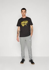 Camiseta de algodón negra con gráfico amarillo que presenta "BVB 09". Pantalones deportivos grises con logo blanco de Puma, combinados con zapatillas negras.