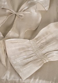 Beige satijnen top met gerimpelde kraag en strikdetail. Heeft een zachte textuur, delicate strikjes en elastische rimpeling bij de taille.
