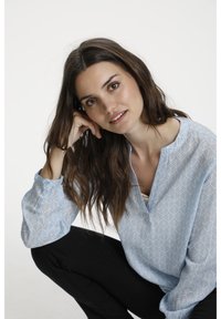 Blouse bleu clair avec un motif de feuilles, un col sans col, et de longues manches légèrement froncées. Associée à un pantalon noir.