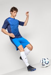 Tricou de fotbal albastru și bleumarin cu design texturat, asortat cu șorturi albastre. Modelul poartă șosete albe și ghete negre, mingea la picior.