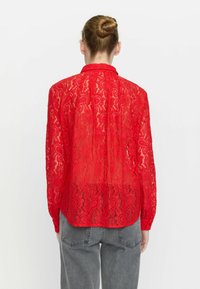 Red lace blouse with long sleeves, a collar, and intricate paisley pattern; semi-transparent with a straight silhouette and buttoned cuffs.