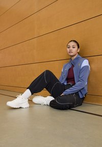 Chaqueta deportiva de color azul claro con una parte superior marrón, pantalones deportivos negros y zapatillas blancas. El conjunto presenta colores contrastantes y detalles de ajuste ceñido.