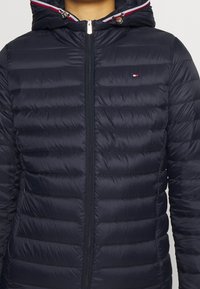 Nærbillede af en person iført en marineblå quiltet jakke med hætte, lynlås foran og et lille rødt, hvidt og blåt logo på brystet.