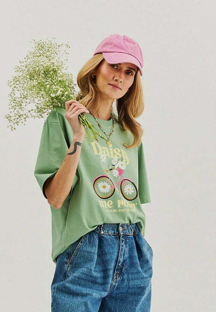 Young woman wearing pink cap, green graphic t-shirt, and blue jeans holding a bouquet of white flowers over her shoulder against a plain background.