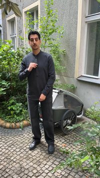 Άνδρας με σκούρο πουκάμισο και παντελόνι στέκεται σε λιθόστρωτο μονοπάτι κοντά σε πράσινα φυτά και δίτροχη ποδηλατική σκεπή δίπλα σε κτίριο.