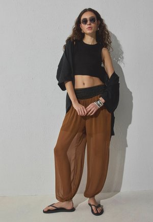 Ung kvinde iført sort ærmeløs crop top, løse brune bukser, oversized sort skjorte, sorte klipklappere, solbriller og sølvarmbånd, stående.