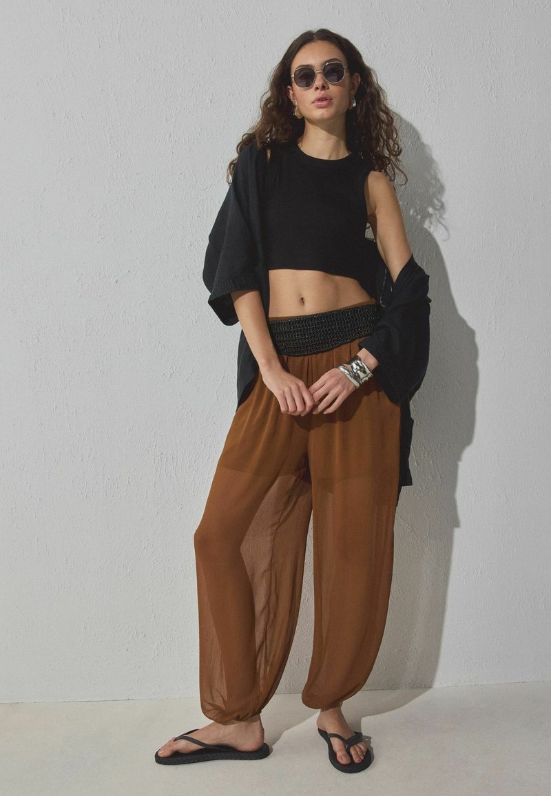 Jeune femme portant un haut court noir sans manches, un pantalon ample marron, une chemise noire oversize, des tongs noires, des lunettes de soleil et des bracelets argentés, debout.