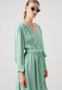 Rochie verde mentă cu decolteu adânc în V, mâneci lungi și talie legată. Materialul neted are un model subtil. Este însoțită de ochelari de soare de culoare deschisă.