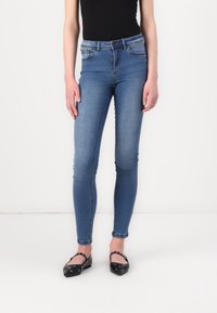 Jeans de denim azul ajustados con una cintura de altura media, cinco bolsillos y una textura suave. Combinados con zapatos planos negros con tachuelas plateadas.