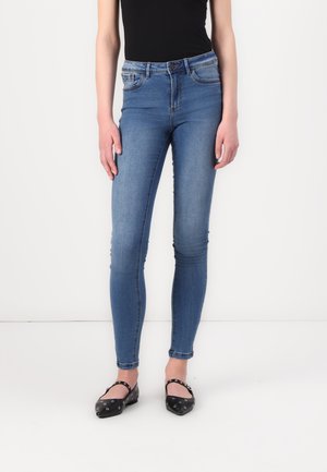 Jeans de denim azul ajustados con una cintura de altura media, cinco bolsillos y una textura suave. Combinados con zapatos planos negros con tachuelas plateadas.