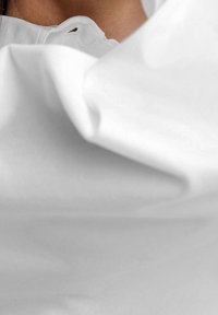 Tissu blanc avec une texture douce et lisse, présentant des plis. Col partiellement visible avec un petit œillet en détail.