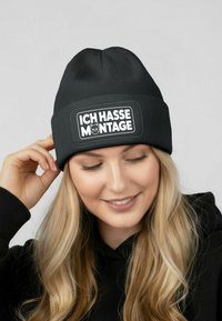 Black ribbed beanie featuring a rectangular patch with the text "ICH HASSE MONTAGE" and a cat illustration. Soft texture with a snug fit.