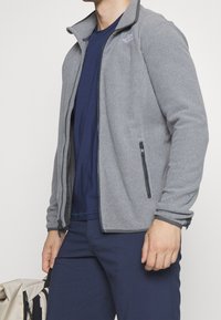 Gray fleece jacket with zippered front, black accents, and side pockets, worn over a navy blue shirt and paired with dark pants.