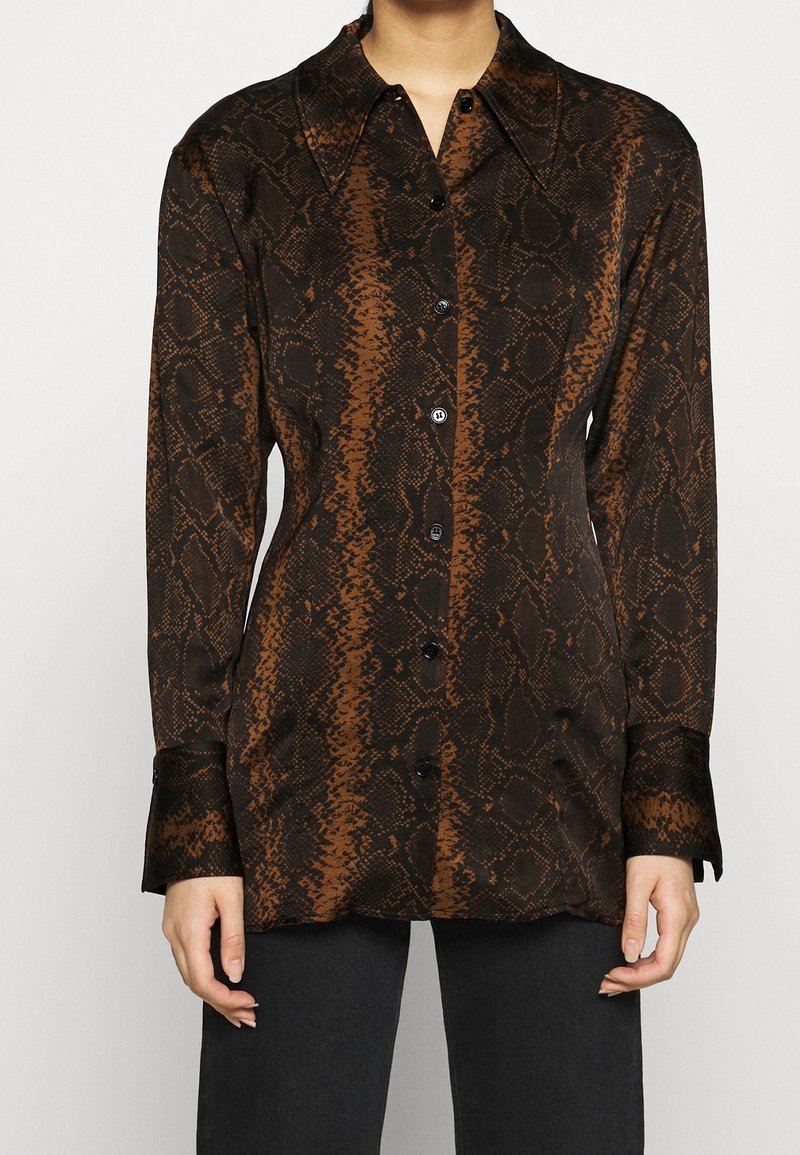Femme portant une chemise à manches longues boutonnée avec un imprimé serpent brun et noir et un col pointu, associée à un pantalon noir.