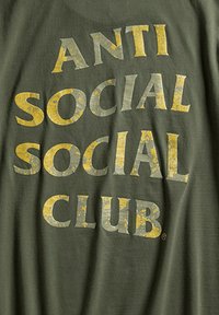 Olivegrzna koszulka z dużą, zdeformowaną żółtą i szaro-typografią z napisem "ANTI SOCIAL SOCIAL CLUB". Miękka tkanina z luźnym krojem.