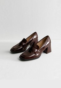 Mocasines de charol marrón con tacón cuadrado, que presentan una puntera redondeada y una correa decorativa en el empeine. Acabado suave y brillante.