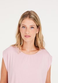 Top rosa chiaro a maniche corte realizzato in tessuto morbido con scollatura rotonda e dettagli plissettati vicino al colletto. Design semplice e liscio.
