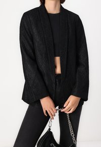 Femme portant un blazer en dentelle florale noire, un crop top noir, un pantalon noir, et tenant un sac à main noir avec une bandoulière chaîne argentée.