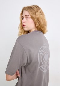 T-shirt grigia a maniche corte con un logo circolare con un design a forma di cuore sulla schiena, realizzata in morbido materiale di cotone.