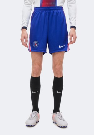 Person iført blå Paris Saint-Germain fodboldshorts, sorte Nike strømper, hvide fodboldstøvler og en hvid langærmet trøje står oprejst.
