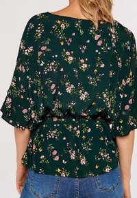 Blouse fleurie avec un fond vert foncé, ornée de fleurs roses, jaunes et blanches, avec des manches amples et une taille ceinturée pour la silhouette.