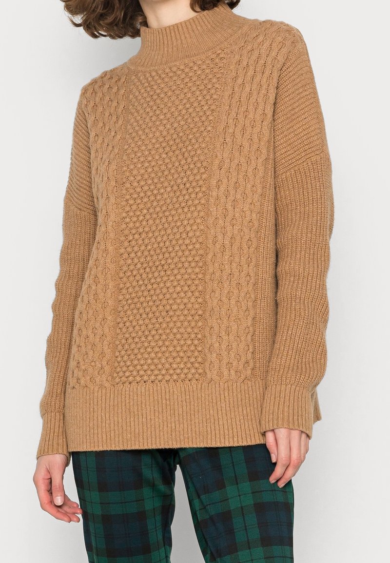 Brązowy sweter dziany z wysokim kołnierzem, z wzorami w warkocze i pomponami, ściągacze na rękawach oraz luźny krój. Noszony na zielone w kratę spodnie.